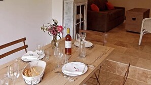 Un coin repas avec une table en bois dressée pour un repas, un vase rempli de fleurs et une bouteille de vin. On aperçoit un canapé marron et un téléviseur au fond.