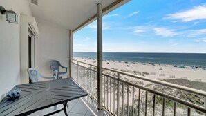 Un balcon avec une table et des chaises, donnant sur une plage où l’on peut voir des gens et des transats.