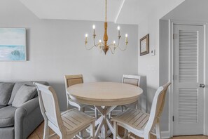 Ein Essbereich mit einem runden Tisch und sechs Stühlen, einer Chandelüster, einem grauen Sofa und einer weißen Tür mit Lamellen.