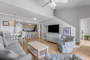 Ein geräumiges Wohnzimmer mit grauem Sofa, einem gemusterten Sessel und einem Holztisch. Ein Fernseher ist an der Wand montiert, und eine Treppe führt zu einer weiteren Ebene.
