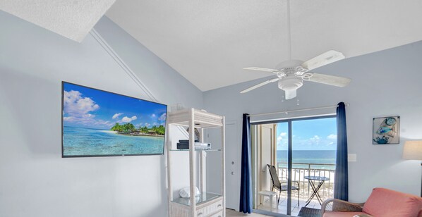 Una stanza con una ventola a soffitto, un'ampia finestra vista mare, un quadro appeso raffigurante una spiaggia e una sedia vicino alla finestra.