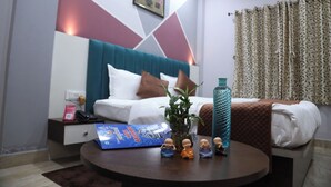 A hotel room with a bed, a bedside table with a book and a plant, a water bottle, and a small Buddha statue collection. - hotel basurivala  (Bhopal)