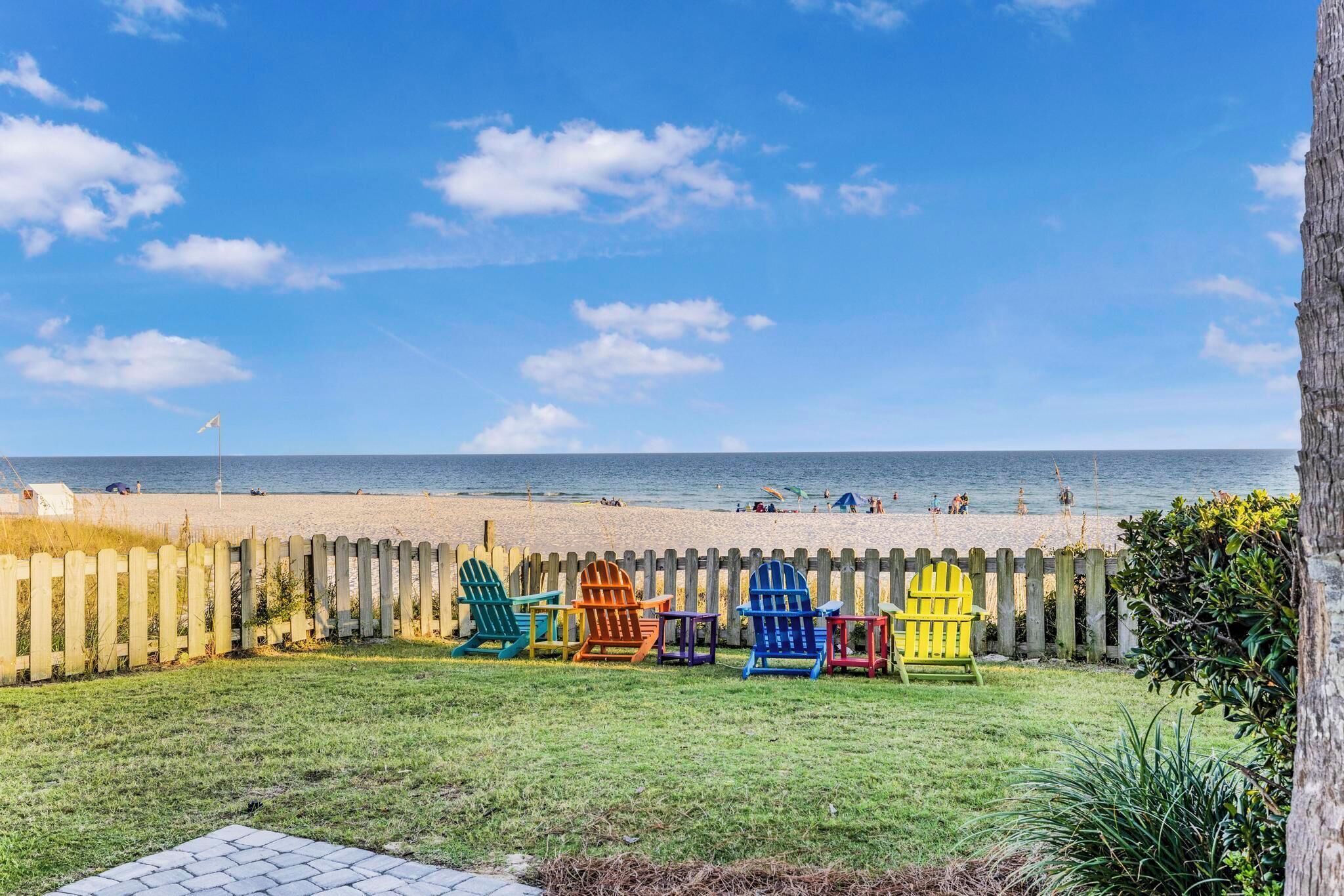 Vista de la playa con una valla de madera y sillas Adirondack de colores en primer plano.