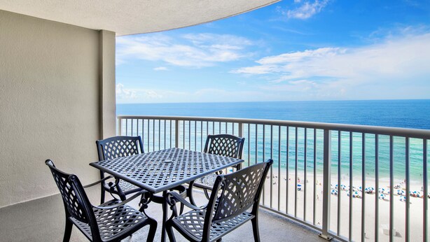 Ein Balkon mit Tisch und Stühlen mit Blick auf einen Strand mit Sonnenschirmen und Menschen.