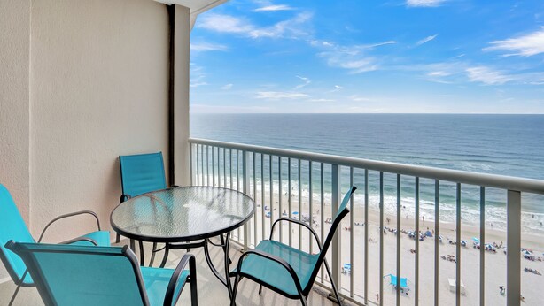 Un balcon avec une table et des chaises en verre, donnant sur une plage parsemée de parasols et fréquentée par des gens.