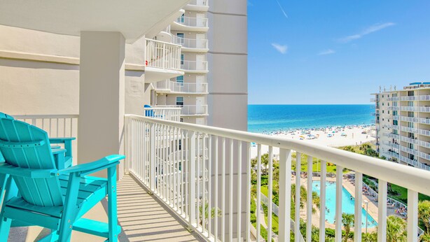 A balcony with turquoise chairs, a white railing, and a view of a beach and pool area.