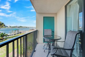 A balcony with a glass table and chairs, a teal door, and a view of the water and distant buildings. - 🌴Wind Drift 114 North | Water Front with Gorgeous Views | Walk to Beach🌴 (Orange Beach)