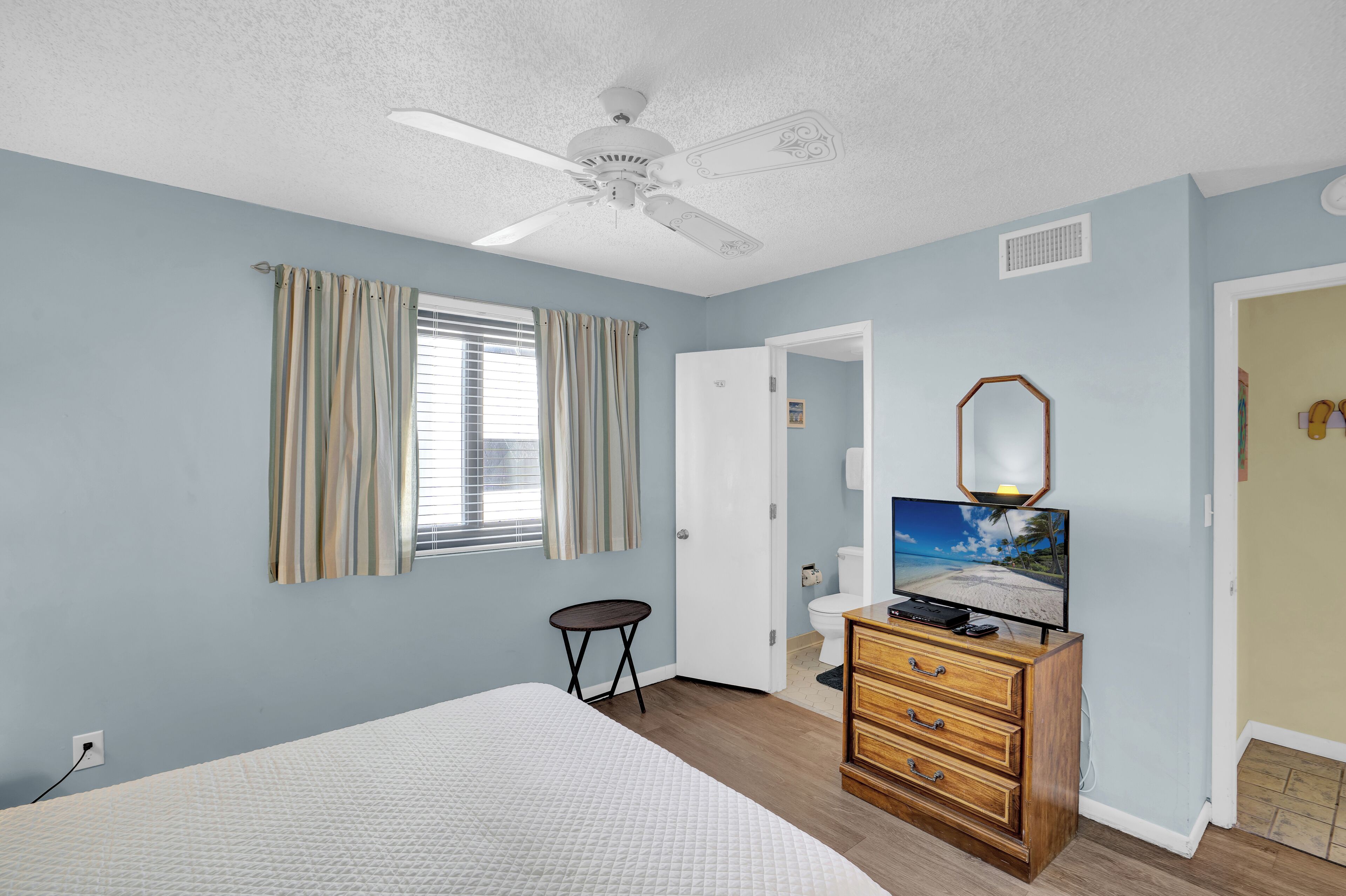 Une chambre à coucher comprenant un lit, un ventilateur de plafond, un téléviseur et une commode en bois.