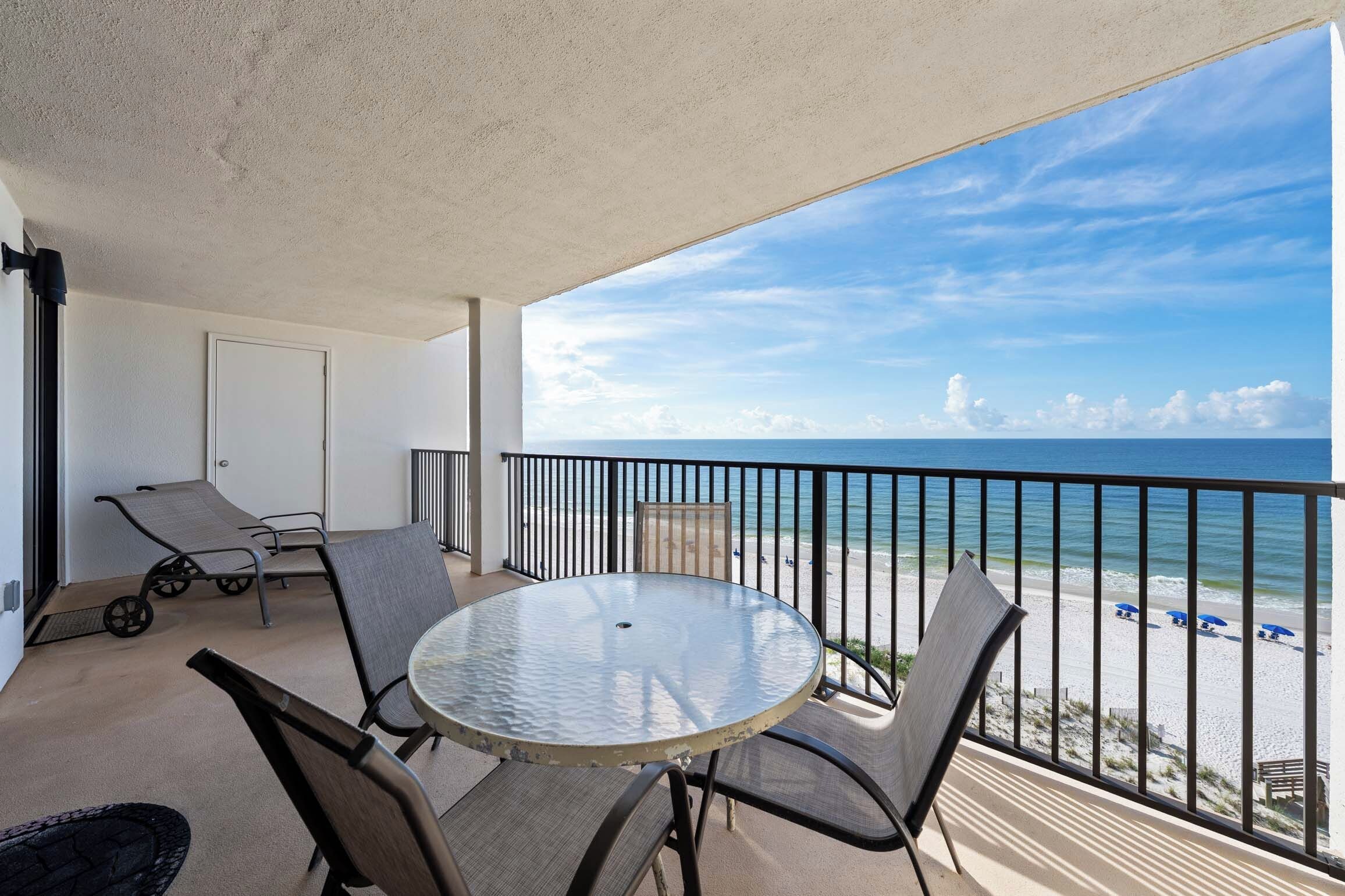 A balcony with a round table and chairs, a view of the beach, and a door to the left.