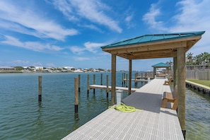 A wooden pier extending into calm water, with a covered seating area and a view of distant buildings.