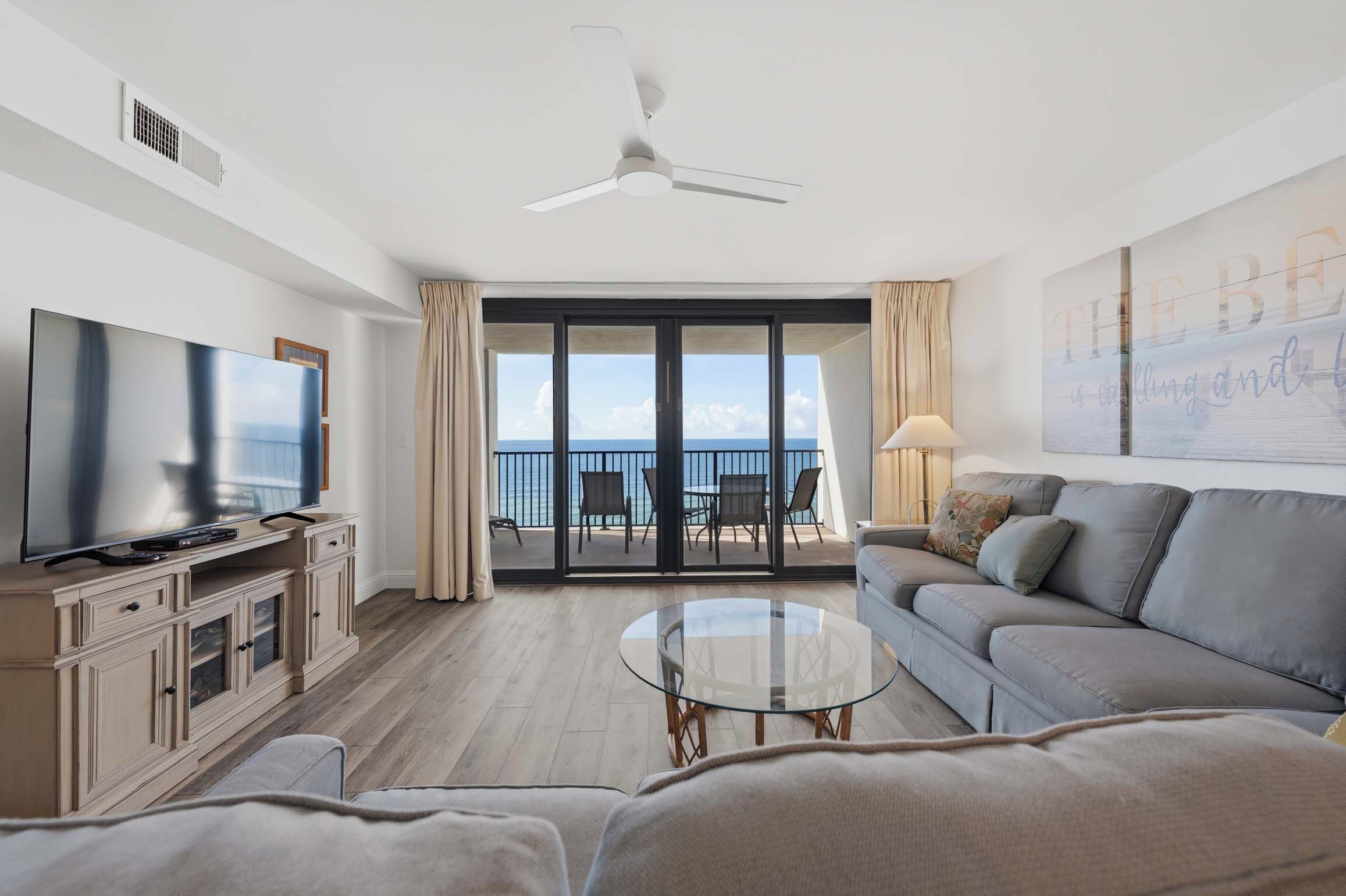 A modern living room with a flat-screen TV, a sofa, a glass coffee table, and a ceiling fan.