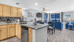 A modern kitchen with wooden cabinets, a built-in dishwasher, and a granite countertop. There is a dining area with blue sofas and a small table.
