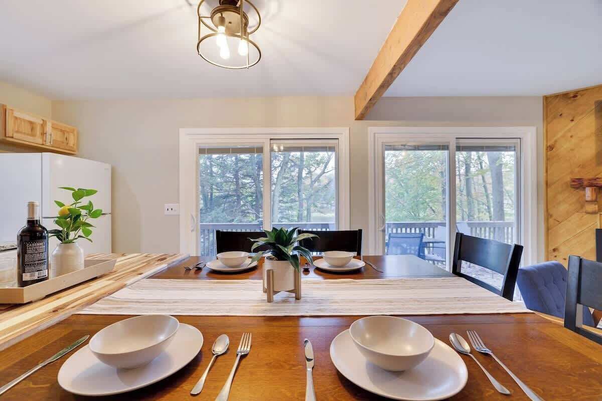 Un coin repas avec une table en bois dressée pour deux personnes, de l’assiette blanche et des couverts en argent. Deux fenêtres offrent une vue sur les arbres.