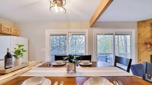 A dining area with a wooden table set for two, white plates, and silverware. There are two windows with a view of trees.
