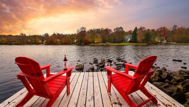 A wooden dock with two red Adirondack chairs, a bottle of wine, and a glass on the dock.