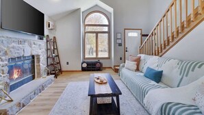 A living room with a stone fireplace, a large TV, a wooden staircase, and a cozy sofa.