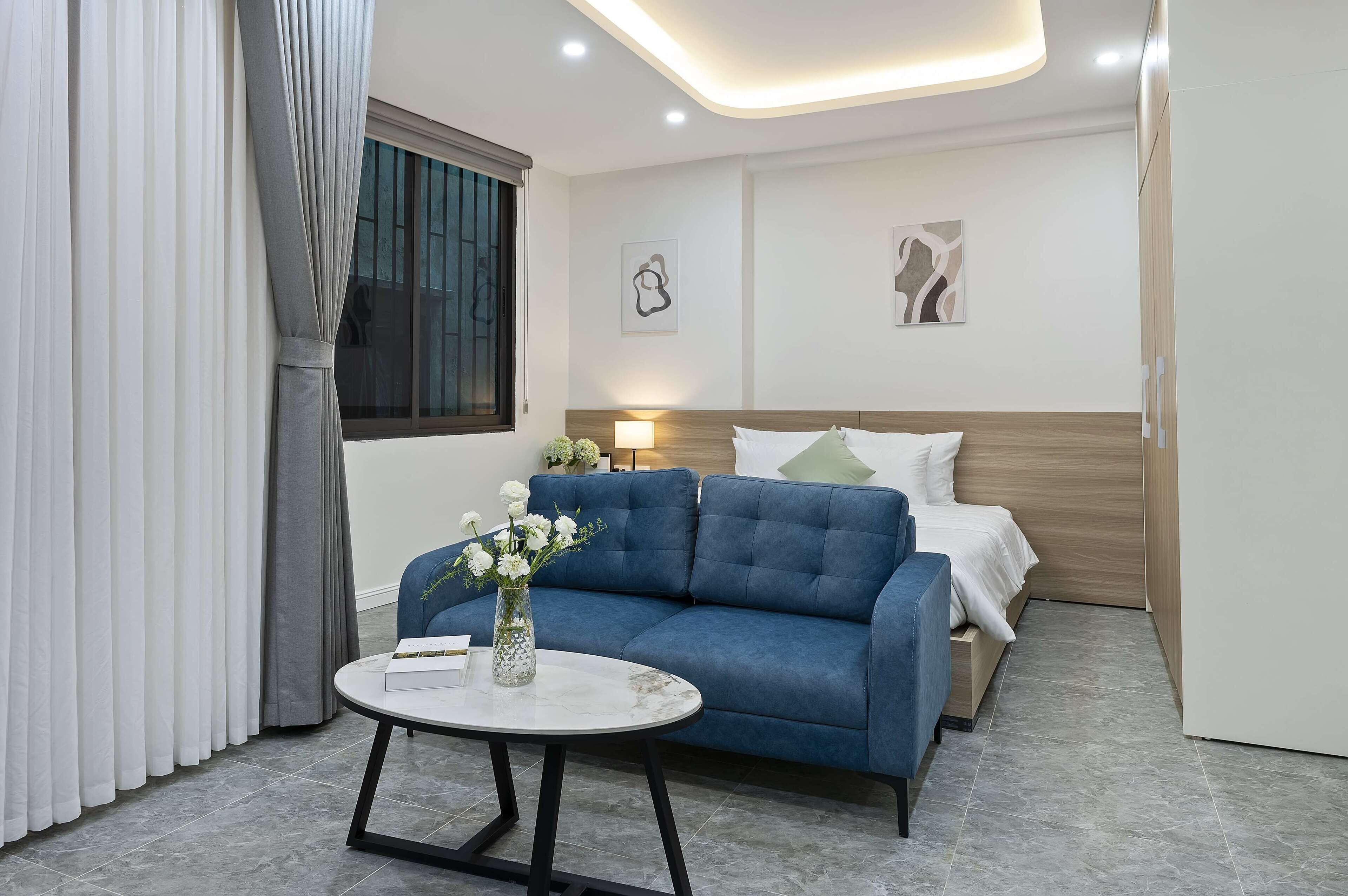 A modern hotel room with a blue sofa, a small round table with a vase of flowers, a bed with white bedding, and two abstract wall art pieces.