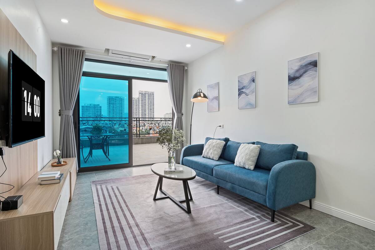A modern living room with a blue sofa, a coffee table, a flat-screen TV, and a view of a balcony with a cityscape.
