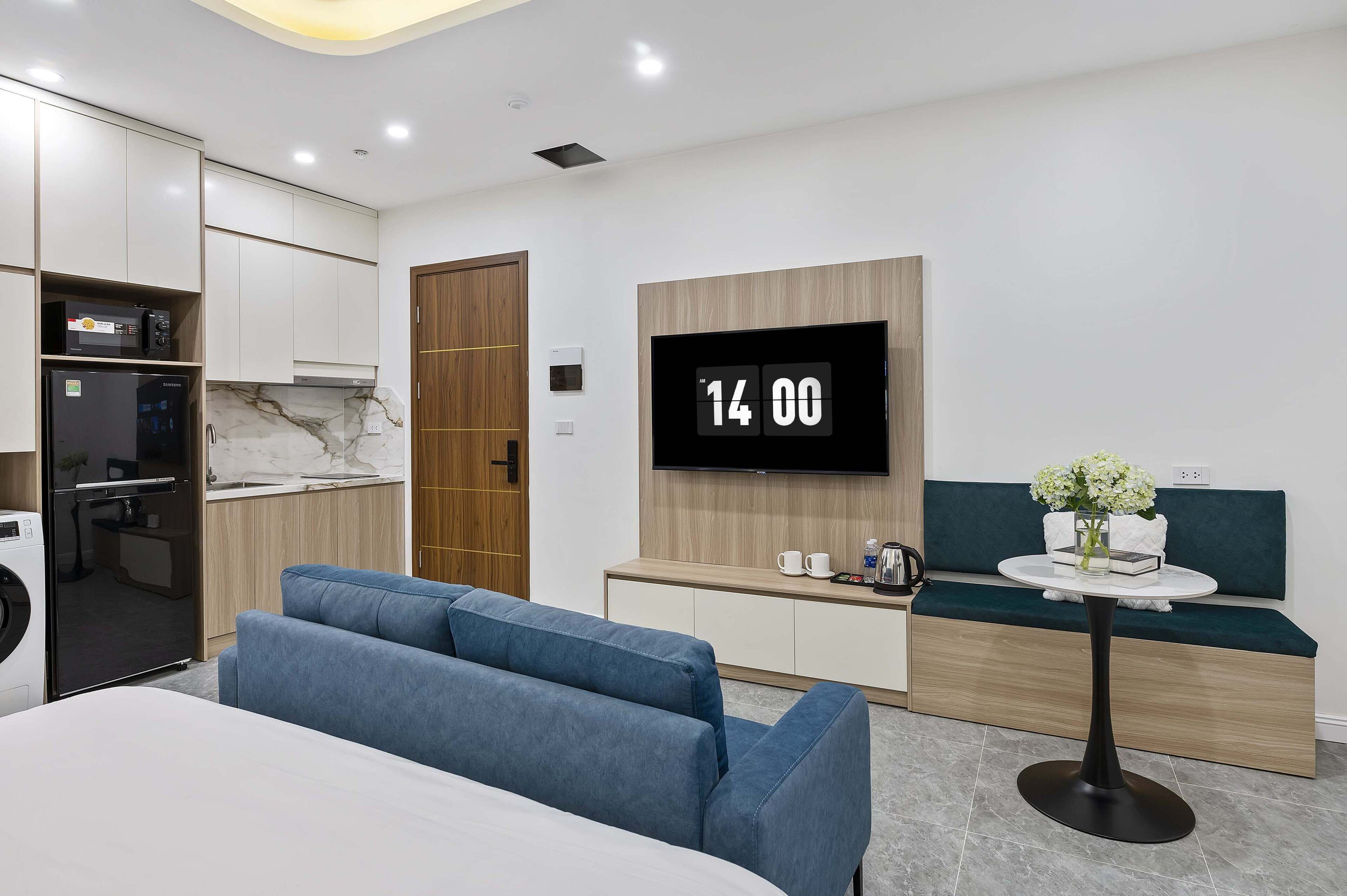 Ein modernes Hotelzimmer mit einem Bett, einer Sitzecke mit blauen Sofas, einem kleinen Tisch mit einer Vase und einem Fernseher, der die Uhrzeit anzeigt.