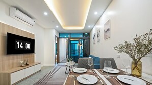 A modern dining area with a table set for two, a flat-screen TV displaying the time, and a view into the living room. - Cosmos Hotel Apartment (Hanoi)