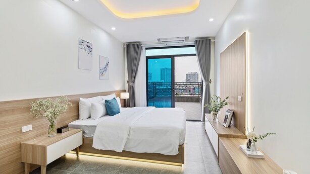 A modern hotel room with a large bed, bedside table, and a view of the city through a glass door. - Cosmos Hotel Apartment (Hanoi)