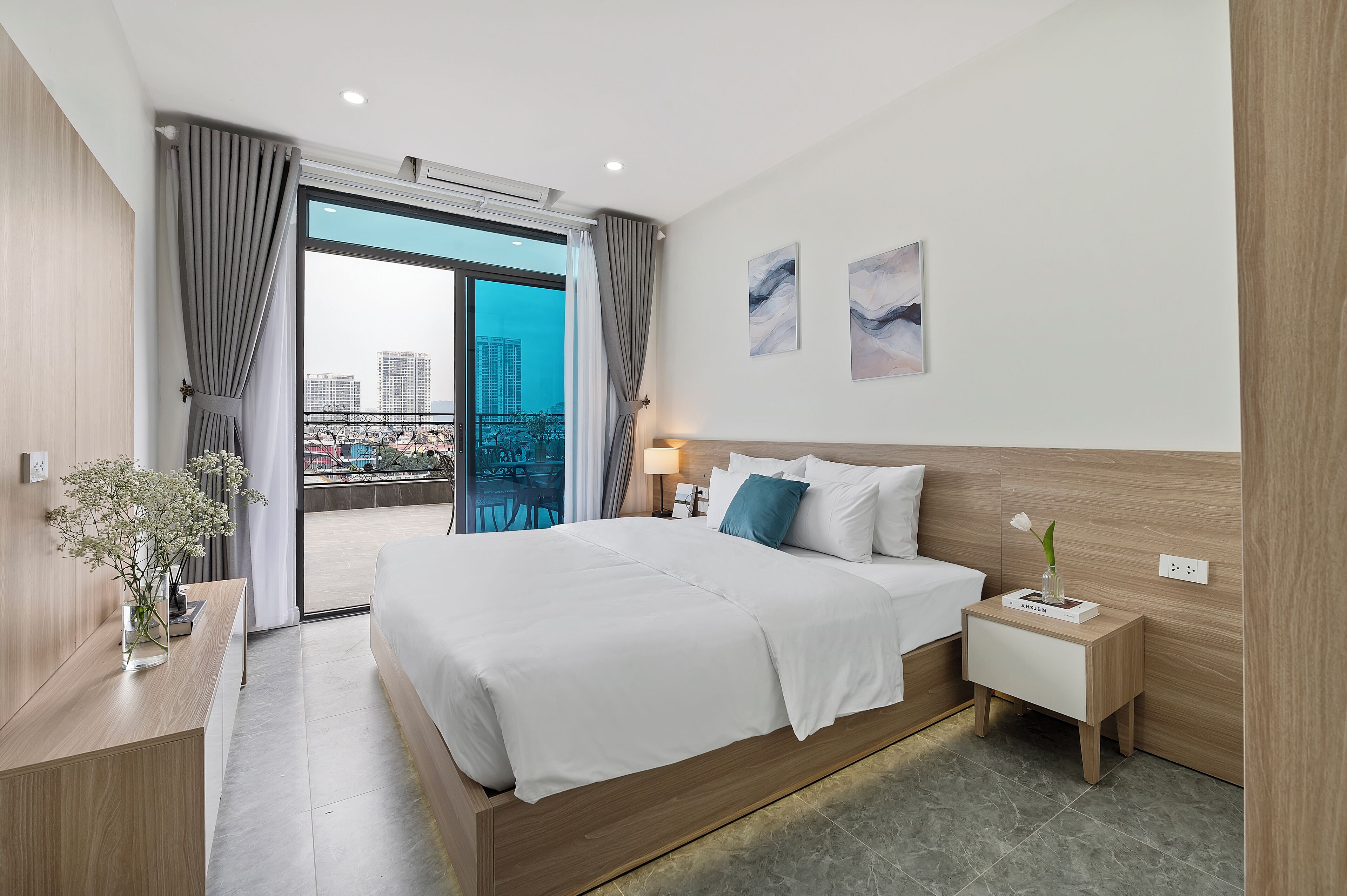Una habitación de hotel moderna con una cama grande, una mesita de noche, un florero con flores y vistas a la ciudad desde un balcón.