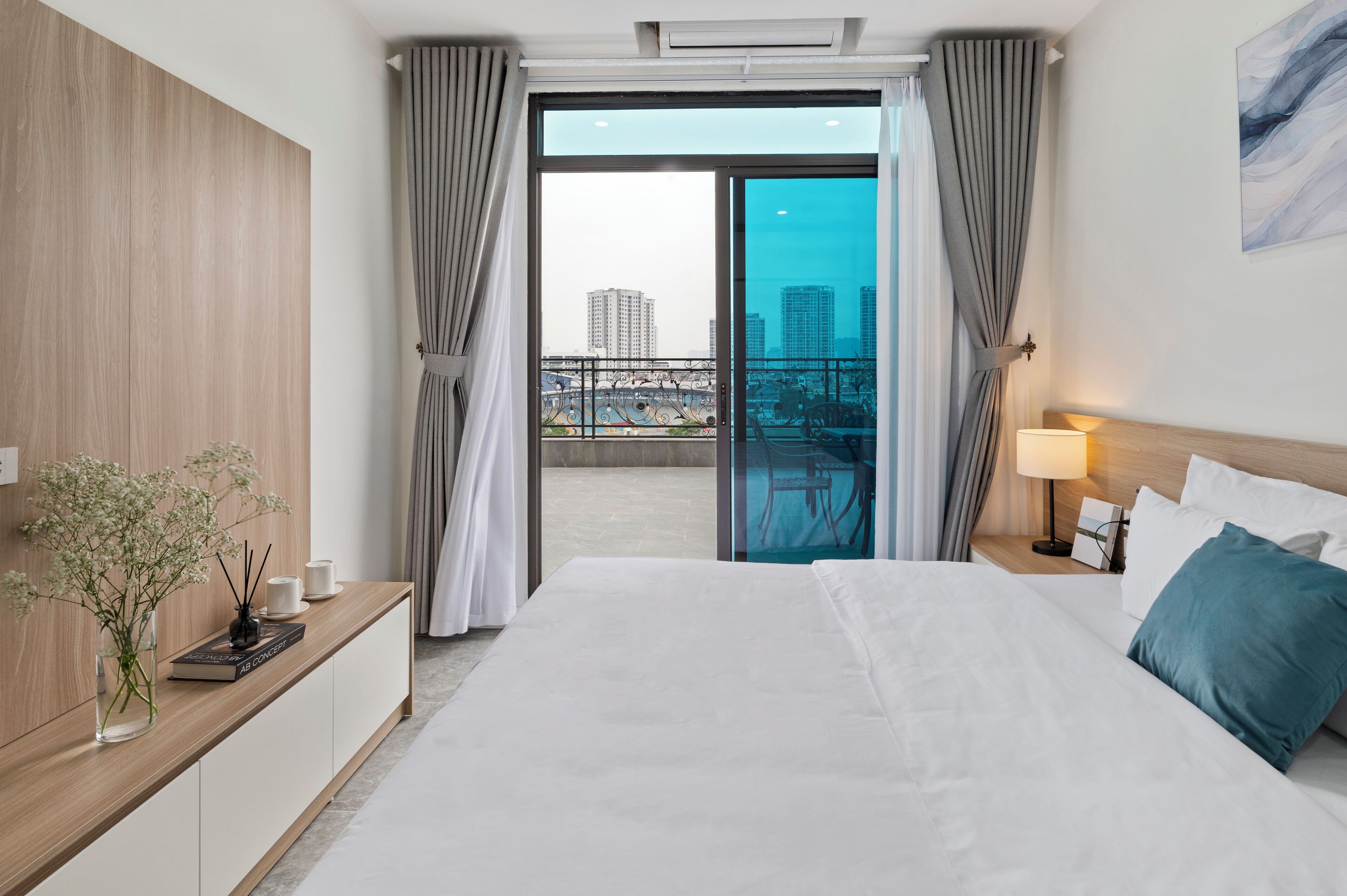 A modern hotel room with a large bed, a bedside table with a lamp, a view of the city through a glass door, and a wooden panel wall.