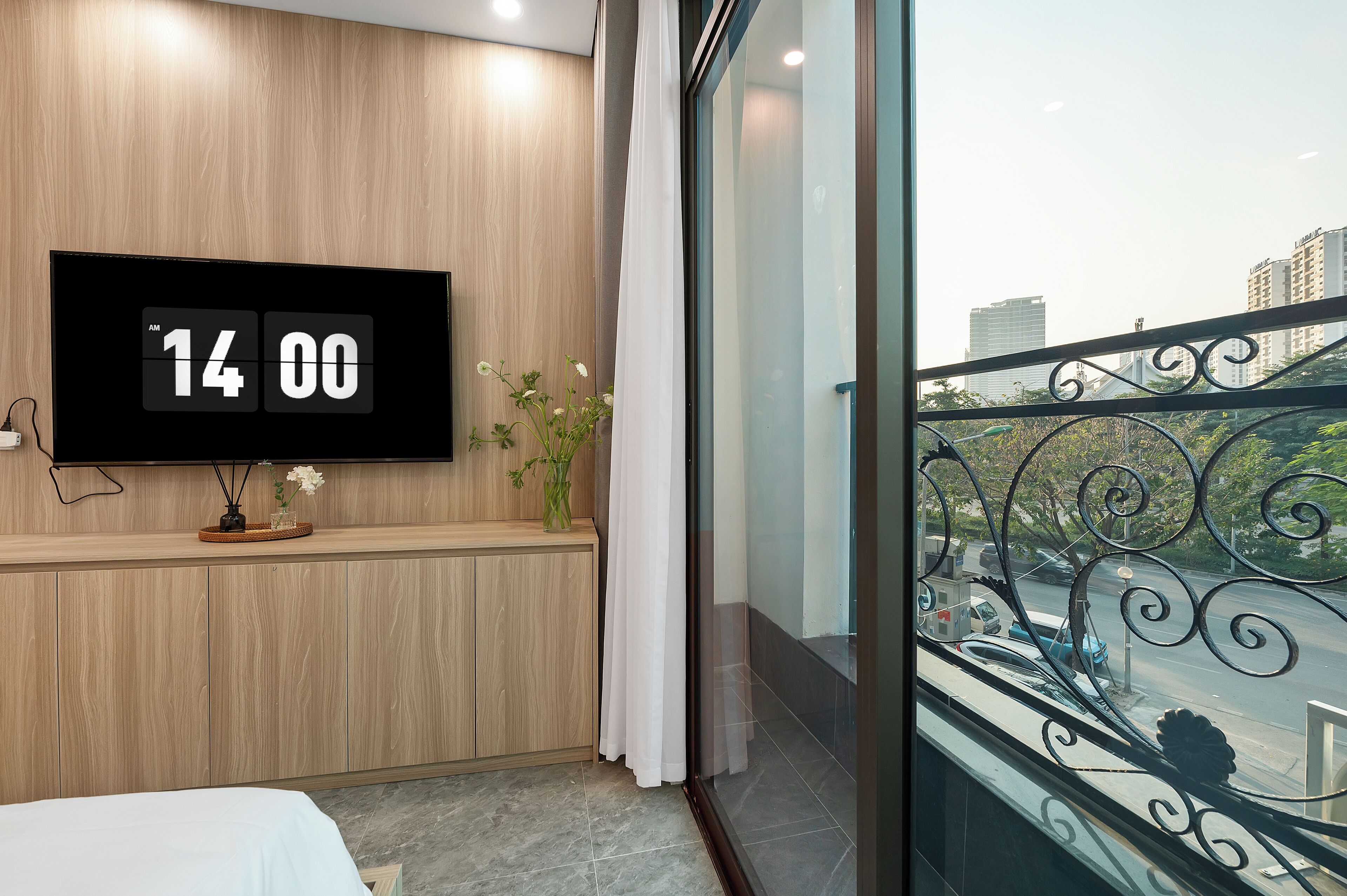 A modern hotel room with a wooden panel wall, a large TV displaying the time, a small plant, and a balcony with a metal railing.