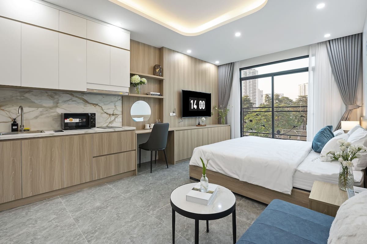 A modern hotel room with a kitchen area, a bed, a TV, and a large window with a city view.