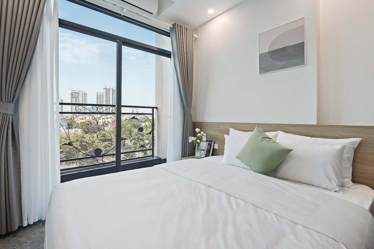A hotel room with a large bed, a view of buildings through a window, and a framed artwork on the wall.