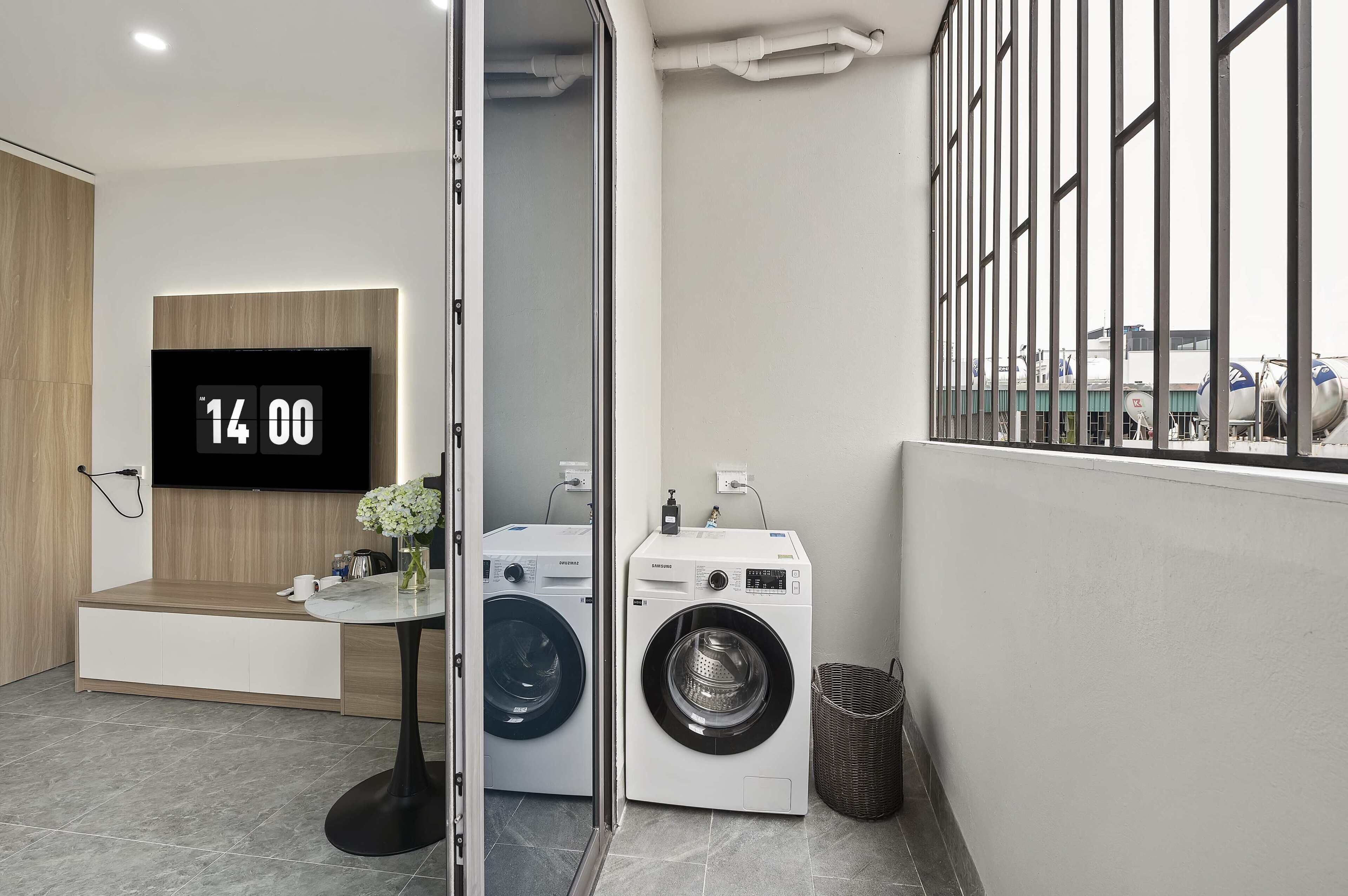 A modern laundry room with two washing machines, a digital clock displaying 14:00, a small table with a vase, and a window with metal bars.