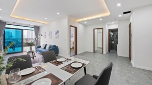 A modern living room with a dining area, large windows, and a view of the city. - Cosmos Hotel Apartment (Hanoi)