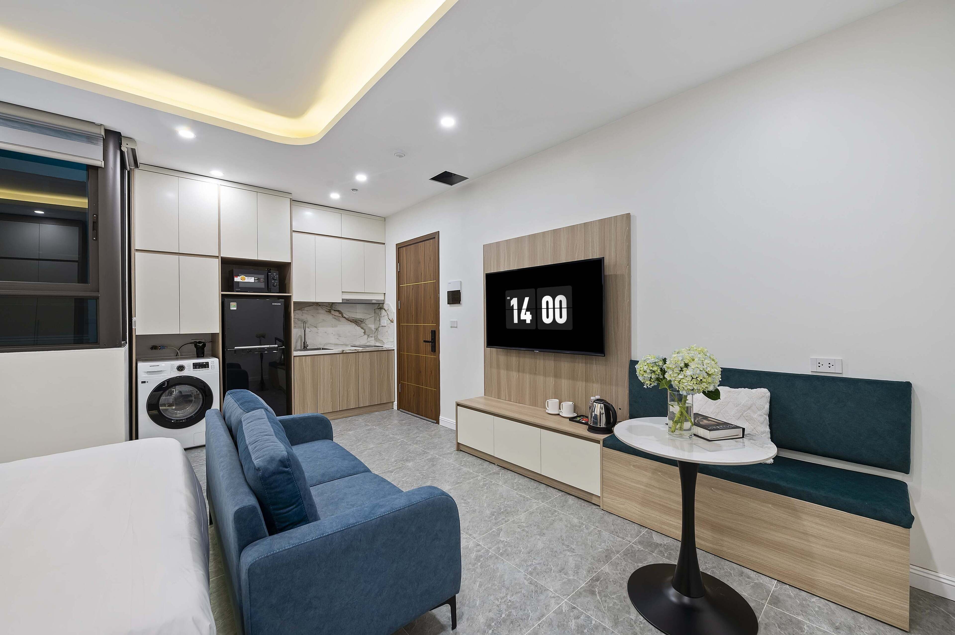 Un espacio habitable compacto con sofá, una mesita con un florero, televisor montado en la pared, lavadora y kitchenette.