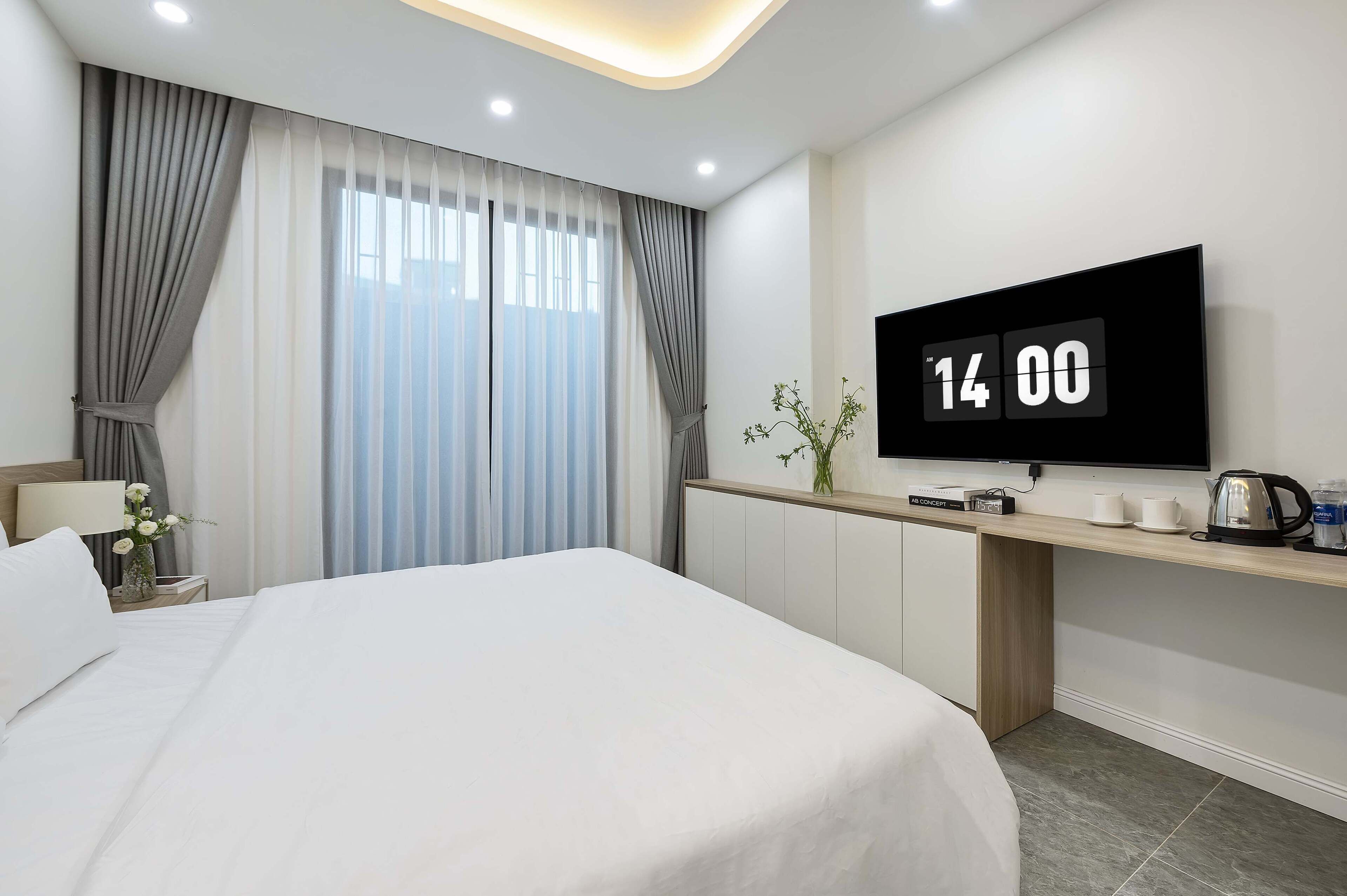 A hotel room with a bed, a TV mounted on the wall, a bedside table with a lamp, and a clock displaying 14:00.
