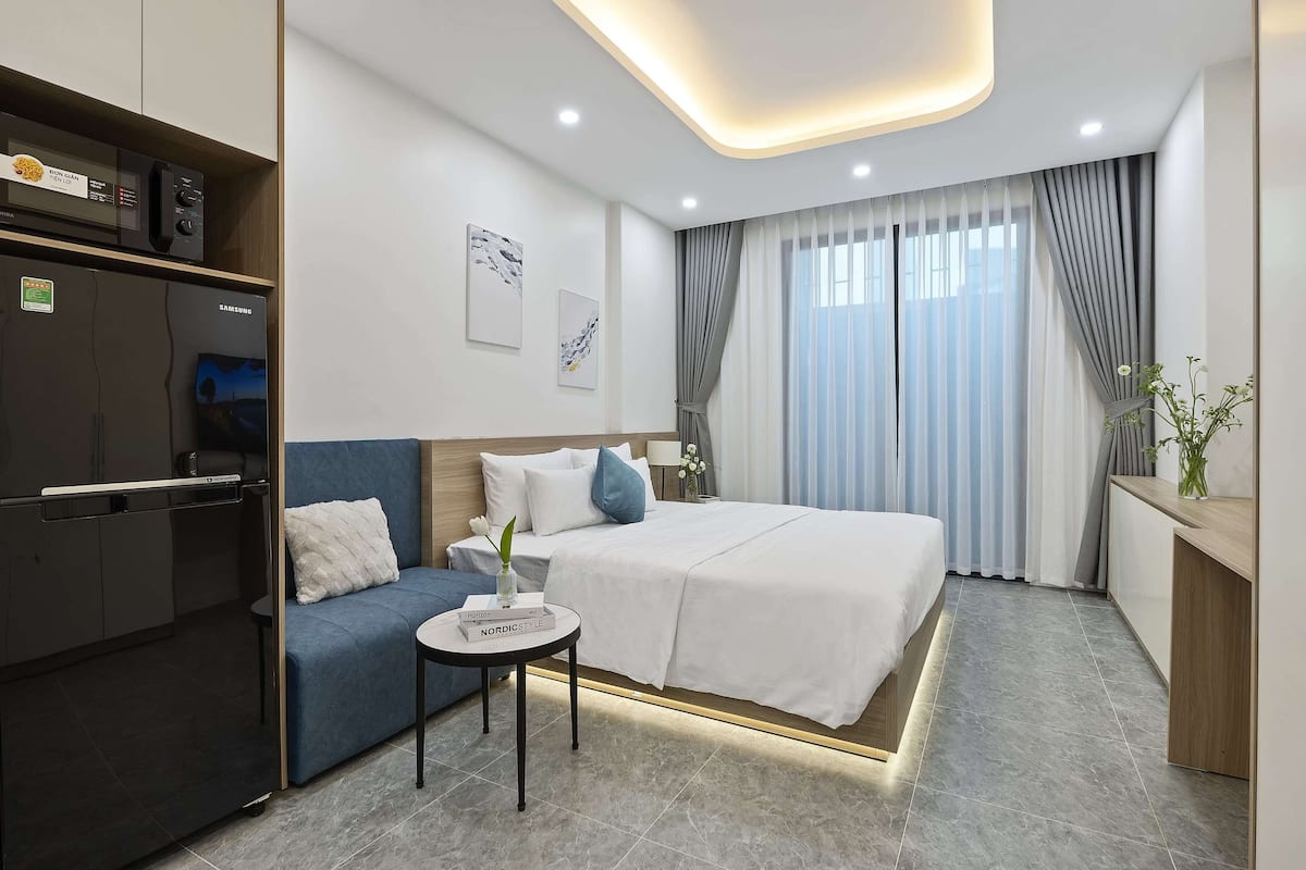 A modern hotel room with a bed, a sofa, a small table, a desk, and a minibar.
