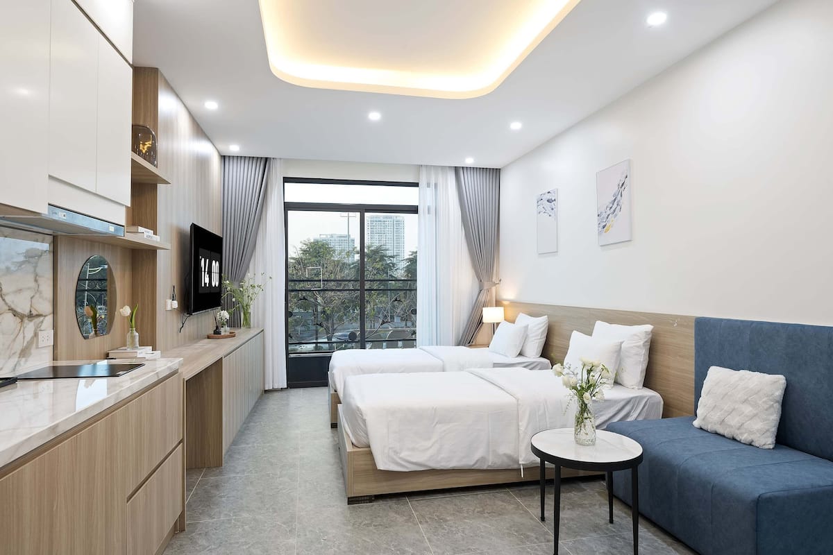 A modern hotel room with a bed, a sofa, a small table, and a kitchen area.