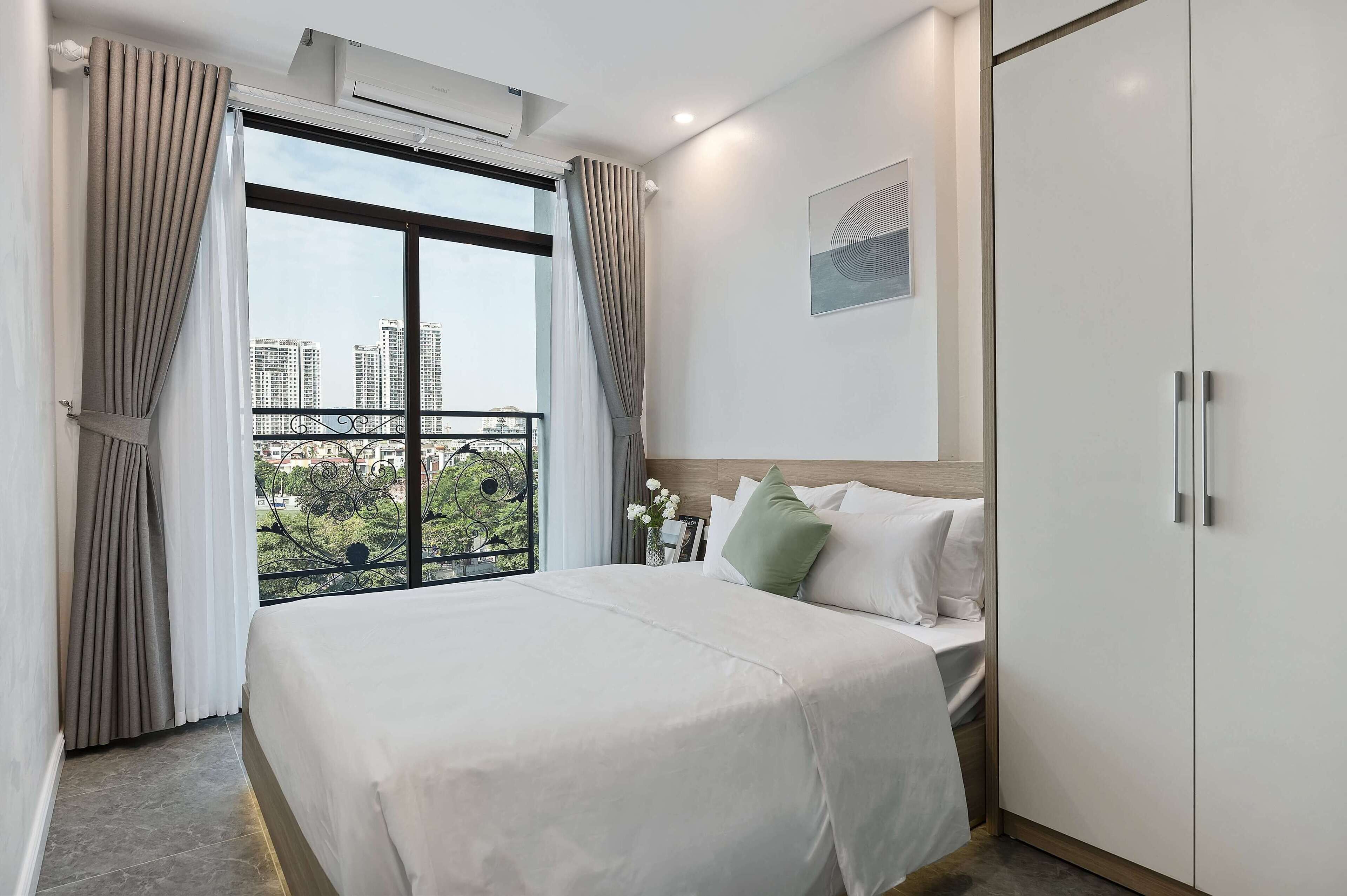A hotel room with a bed, a wardrobe, a window with curtains, and a view of buildings and greenery.