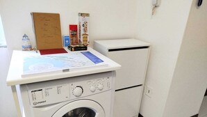 A compact laundry room with a front-loading washing machine, a small refrigerator, and a shelf with various items including a map and a box of tissues. - Historic apartment on Avenue (Trieste)