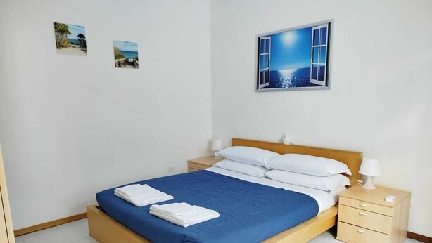 A neatly made bed with blue bedding and white pillows, a wooden headboard, a bedside table with a lamp, and a wall-mounted picture of a beach scene. - Historic apartment on Avenue (Trieste)