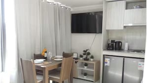 Et kompakt værelse med et spisebord, stole, et lille tekøkken med et køleskab og et TV ophængt på væggen.