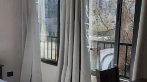 Et hotelværelse med en stor seng, en træstol og udsigt til høje bygninger gennem lette gardiner.