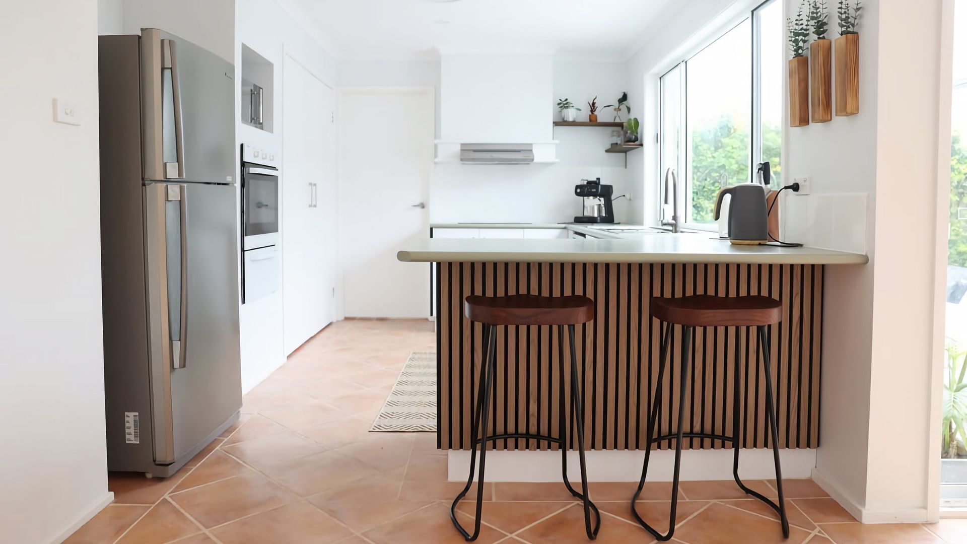Una cocina moderna con refrigerador, horno y microondas integrados, una isla de madera con taburetes altos y vistas a zonas verdes.