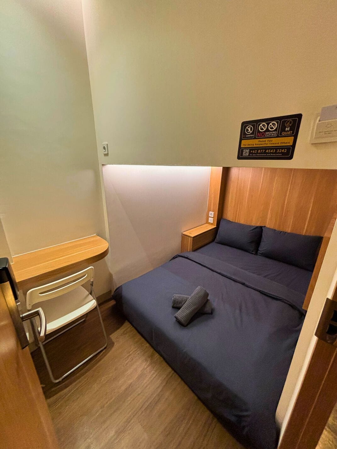 A compact hotel room with a single bed, a bedside table, a small desk with a chair, and a wall-mounted sign with rules.