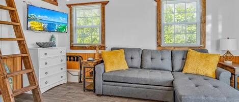 A cozy living room with a grey sofa, yellow cushions, a wooden ladder, and a television on the wall.