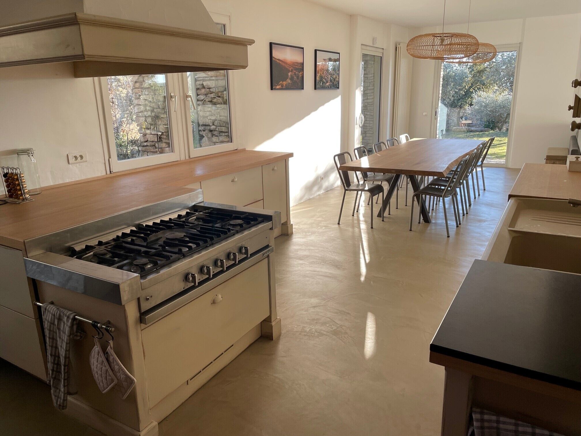 A modern kitchen with a central island, a dining area with a large table and chairs, and a view of the outdoors through large windows.