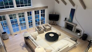 Un salon spacieux, doté d’un grand canapé, d’une table basse et d’un téléviseur posé sur un meuble. On peut apercevoir une cheminée et un balcon à travers de grandes fenêtres.