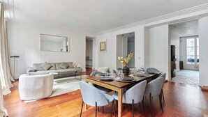 A modern dining area with a wooden table set for four, surrounded by grey upholstered chairs. A grey sofa and a white pouf are visible in the background.