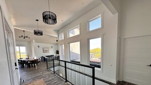 Un interior espacioso con techo alto, ventanas amplias, una escalera con barandilla de vidrio y una zona de comedor con una mesa larga y sillas.
