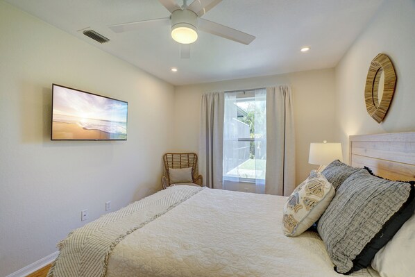 Un dormitorio con cama, ventilador de techo, ventana con cortinas, un cuadro en la pared y una mesita de noche con lámpara.