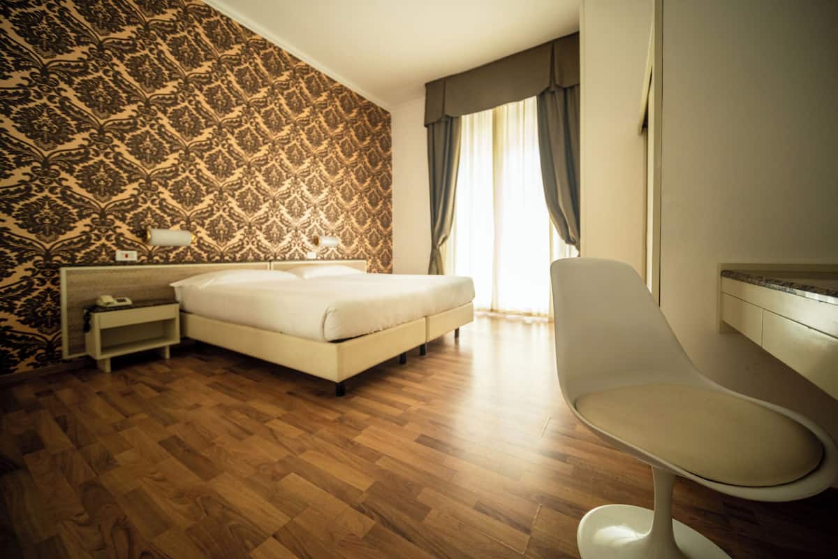 A hotel room with a patterned wallpaper, a bed, a bedside table, a chair, and a large window with curtains.